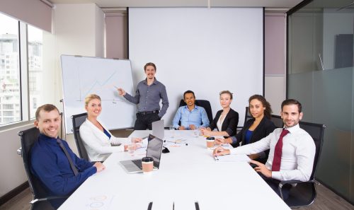 Young businessman standing at whiteboard and pointing to graph with business people sitting at office desk and smiling, looking at camera